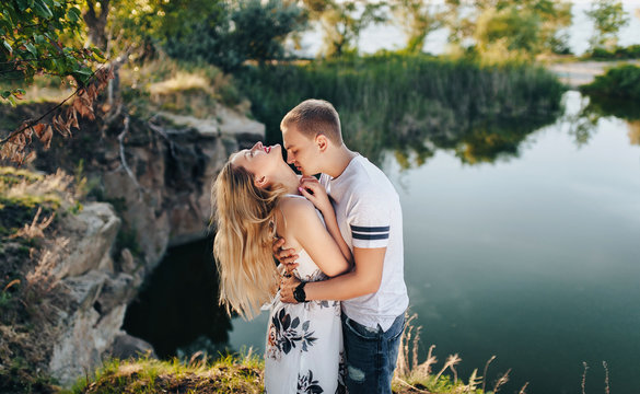 Stylish guy and a young girl hugging against the backdrop of nature, the lake and the sunset with backlight. Closeup portrait of beautiful newlyweds. Kiss on the neck.