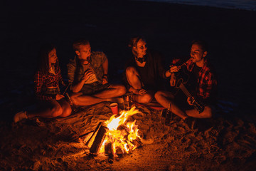 Camp on the beach. Group of young friends having picnic with bonfire. Man is playing guitar, girl is feeding him with sausage
