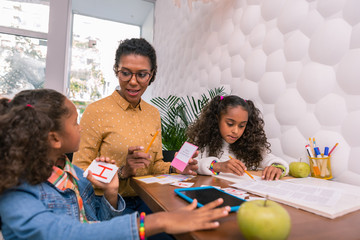 About letters. Attentive pleasant nursery teacher wearing glasses telling cute curly preschoolers about letters
