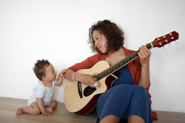 Picture of cute barefooted one year old infant boy sitting on floor with his positive young mom who...