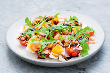 Appetizing salad of cherry tomatoes, eggs, boiled shrimps, arugula, sesame seeds and balsamic sauce in a plate on the table