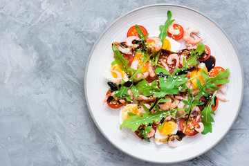 Appetizing salad of cherry tomatoes, eggs, boiled shrimps, arugula, sesame seeds and balsamic sauce in a plate on the table. Top view