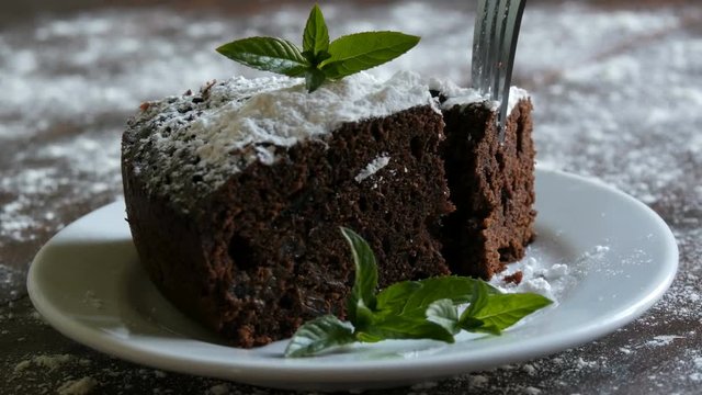 Homemade baked chocolate brownie cake muffled with powdered sugar on a white plate decorated with mint leaves. Fork breaks off piece of brownie pie from the plate