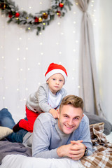 Happy family, father and his son, in the morning in bedroom decorated for Christmas. Boy in Santa's hat sitting on his father. New Year's and Christmas theme. Holiday mood