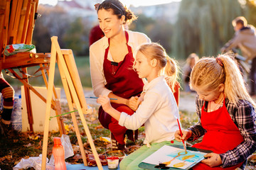 Obraz na płótnie Canvas Young smiling female teacher explaining how drawing for girls paitners outdoor in the park. Open air activity for school age children concept. Real people models.
