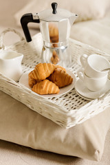 Breakfast in bed. On a white wicker tray there is a coffee maker, coffee white cups and croissants.