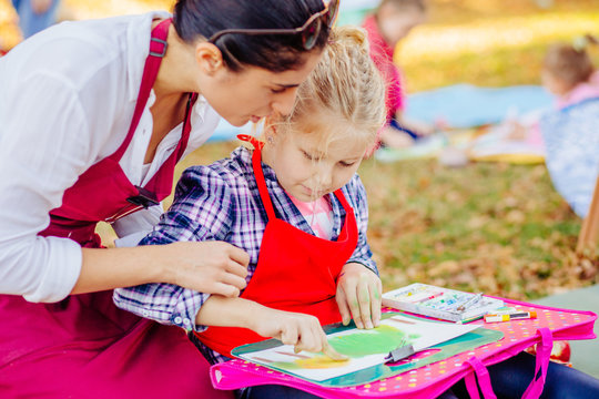 Young female teacher and happy girl in red aprons are painting outdoor in the park. Open air activity for school age children concept.