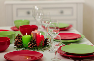 Merry Christmas and Happy New Year! Тable setting festive decor - green and red dishes, candles and fir cones. Knitted decor - Santa Claus knitted hats on the bottle with wine.