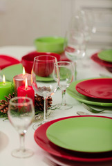 Merry Christmas and Happy New Year! Тable setting festive decor - green and red dishes, candles and fir cones. Knitted decor - Santa Claus knitted hats on the bottle with wine.