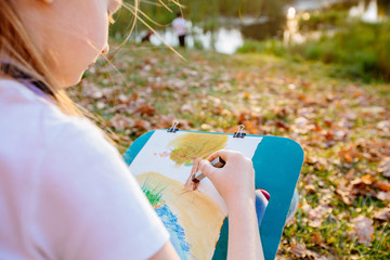Teenager school girl drawing beautiful picture outdoor in the park at sunset. Open air activity for school age children concept. Close up