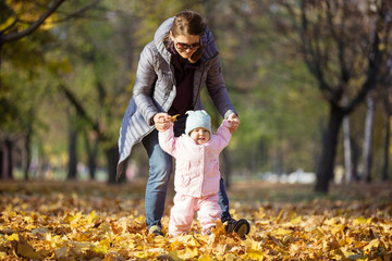 Mother supporting baby daughter and helping her make first steps. Mom and daughter on walk in autumn park.