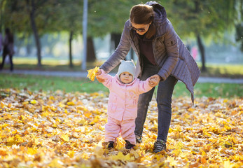 Mother supporting baby daughter and helping her make first steps in autumn park