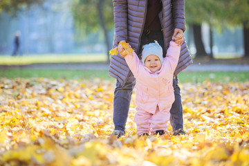 Mother supporting baby daughter and helping her make first steps in autumn park