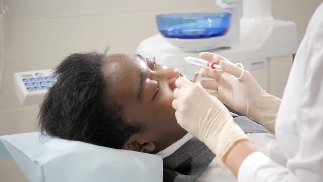 The dentist puts a shot of freezing novocaine in the jaw. Young African American male patient at chair at dental clinic. Medicine, health, stomatology concept.