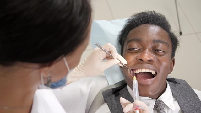 The dentist puts a shot of freezing novocaine in the jaw. Young African American male patient at chair at dental clinic. Medicine, health, stomatology concept.