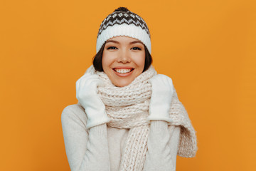 Woman portrait. Accessories. Warmness. Asian girl in a white scarf, cap and gloves is smiling, on an orange background