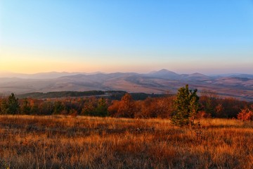 Fototapeta na wymiar A scenic panoramic view of autumn trees, grass, fields and mountains during sunset at golden hour