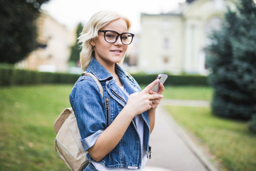 Fototapeta premium Happy woman in eyesglasses smiling and walking in the street using a smartphone and looking at camera