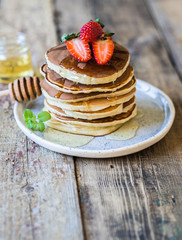 Pile of pancakes with honey and strawberries on a white plate on a wooden table