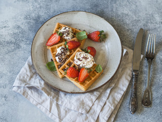 Belgian waffles with cream, chocolate and strawberries on a gray plate on a gray rustic background.