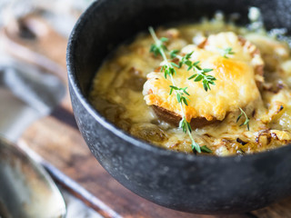 Traditional french onion soup with toast, cheese and fresh thyme in a black bowl on a wooden brown board on a rustic wooden background