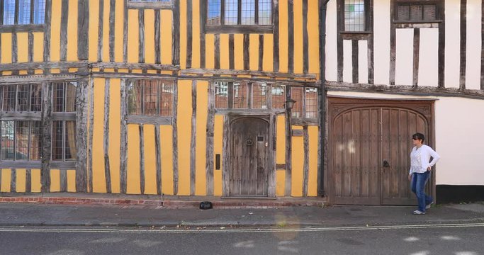 Lavenham England Crooked house woman walks. Lavenham village, civil parish in Suffolk, England. 15th century church, half timbered crooked unlevel medieval cottages and business.