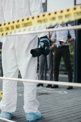 Fototapeta na wymiar cropped view of criminologist in protective suit and latex gloves with camera at crime scene
