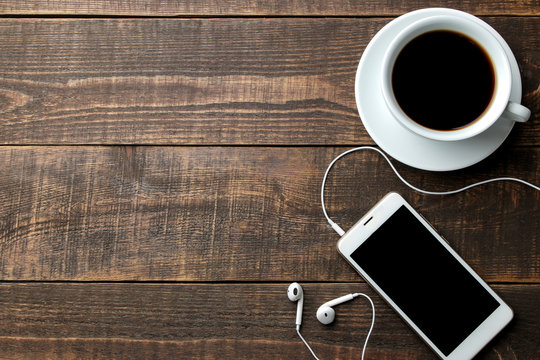 Smartphone with headphones and a cup with coffee on a brown wooden table. view from above