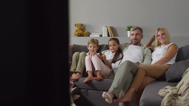 Beautiful mother, father, little kids and grandparents sitting together on comfortable sofa in the living room and watching TV