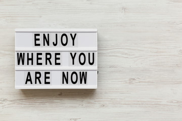'Enjoy where you are now' words on modern board over white wooden background, overhead. Copy space.
