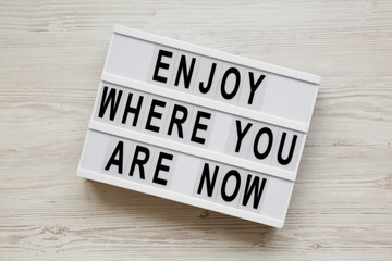 'Enjoy where you are now' words on modern board over white wooden surface, overhead view. Flat lay, from above.