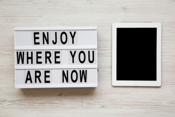 'Enjoy where you are now' words on lightbox, tablet with blank screen over white wooden background, overhead view. Top view, flat lay, from above.