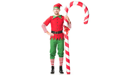 smiling man in christmas elf costume with hand on hips standing near big candy cane isolated on white - obrazy, fototapety, plakaty