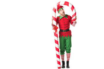 surprised man in christmas elf costume standing under big candy cane isolated on white