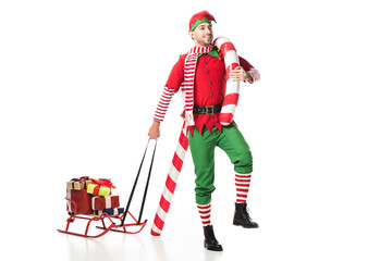 Fototapeta premium smiling man in christmas elf costume carrying sleigh with presents and big candy cane isolated on white