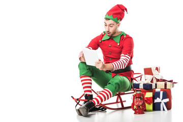 surprised man in christmas elf costume sitting on sleigh near pile of presents and looking at wishlist isolated on white