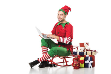 smiling man in christmas elf costume sitting on sleigh near pile of gifts and using laptop isolated...