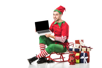 man in christmas elf costume sitting on sleigh and holding laptop with blank screen isolated on...