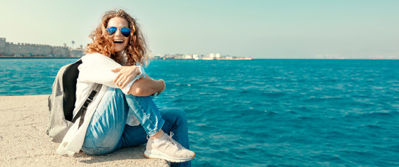 Happy beautiful young woman in a white shirt is sitting on the pier against the blue sea and sky,...