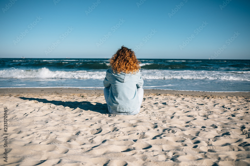 Wall mural young woman sitting on sandy beach looking at the waves