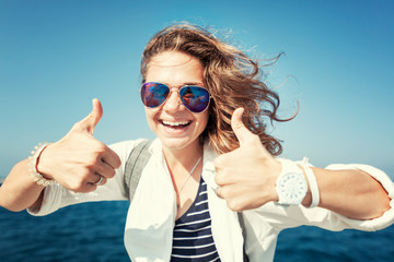 Happy beautiful young woman in a white shirt is sitting on the pier against the blue sea and sky,...