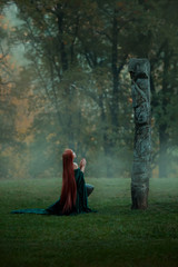 womderful young girl lives in a misty forest, comes to a clearing on a hill to a deity for prayer, a sorceress with fiery red hair, kneels in a long green emerald velvet dress, brave at heart