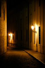 Old lanterns illuminating a dark alleyway medieval street at night in Prague, Czech Republic. Low key photo with brown yellow tones from the lanterns as single light sources against the dark shadows - 233172318