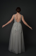 Fototapeta premium full length portrait of brunette girl wearing beautiful long gown, standing pose with background to the camera on grey studio background.
