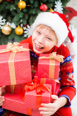 Closeup portrait of cute white kid wearing red santa hat and holding many present boxes in hands. Happy Christmas holiday concept. Vertical color photography.