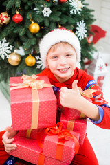 Fototapeta na wymiar Portrait of cute white kid with thumb up. Boy in red santa hat holding stack of holiday Christmas presents. Vertical color photography.