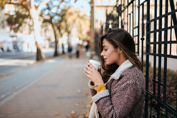 Young stylish woman drinking hot drink in autumn city street.