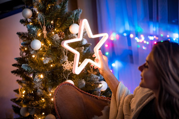 Cheerful young woman sitting next to christmas tree and holding glowing LED star in dark cozy room at xmas eve
