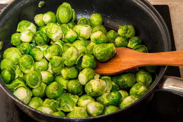 Small green cabbages of Brussels sprouts are stirred with a spatula in a frying pan. Kitchen top plate and stove on the background.