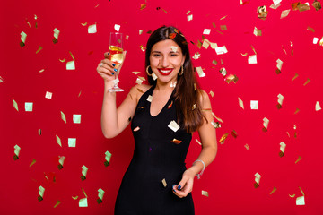 Cheerful young woman is stretching out her hands while confetti falling on her. Beautiful stylish woman in holiday outfit holding champagne against the red background of confetti